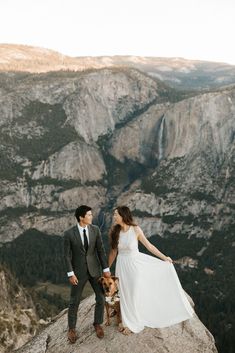 a man and woman standing on top of a mountain holding hands with a dog in front of them