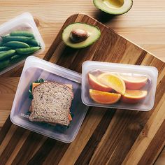 two plastic containers filled with food sitting on top of a wooden cutting board next to an avocado