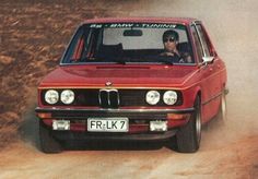 a red car driving down a dirt road next to a person in the driver's seat