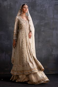 a woman in a white and gold bridal gown, standing against a gray background