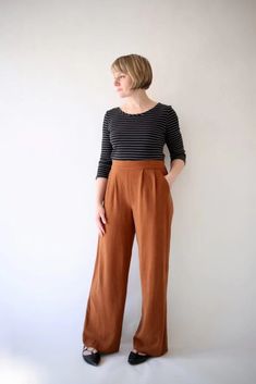 a woman standing in front of a white wall wearing brown pants and a striped shirt