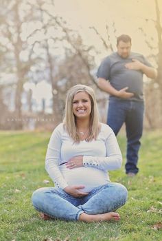 a pregnant woman sitting in the grass with her husband standing behind her and looking into the camera