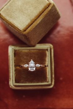 an engagement ring sits in a velvet box on top of a red cloth covered cushion