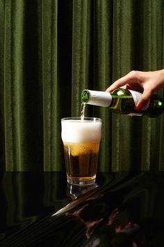 a person pouring beer into a glass on top of a table next to a green curtain