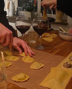 two people are making ravioli on a wooden table with wine glasses and other dishes in the background