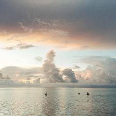 two people riding surfboards in the ocean under a sky filled with clouds at sunset