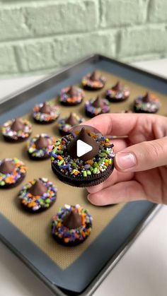 a person is holding a chocolate covered donut in front of a tray of sprinkles