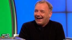 an older man laughing while sitting in front of a tv set with green screen behind him