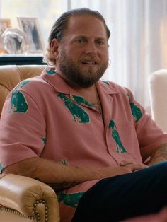 a man sitting in a chair with his arms crossed and looking at the camera while wearing a pink shirt