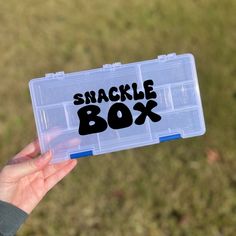 a person holding up a small plastic box with the words snackle box written on it