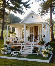 a small white house with flowers and potted plants on the front porch is surrounded by pine trees