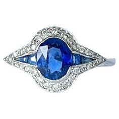 This unusual cluster ring holds sapphires and diamonds around the edges. The sapphire is bright blue in colour. The sapphire total is 1.15 carat and the diamonds total 30pts. Modelled in 18carat white gold. Ring Size: P 1/2 or 7 3/4 Cluster Dimensions: 17x11mm Weight: 3g Blue Oval Sapphire Ring In Art Deco Style, Art Deco Blue Oval Sapphire Ring, Vintage Blue Sapphire Ring With Diamond Accents, Vintage Blue Sapphire Ring With Halo Setting, Art Deco Blue Diamond Ring With Center Stone, Blue Sapphire Ring With Rose Cut Diamonds, Blue Art Deco Diamond Ring With Center Stone, Vintage Blue Cluster Ring With Center Stone, Art Deco Blue Diamond Ring With Rose Cut