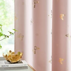 a pink curtain with gold butterflies on it next to a book and potted plant