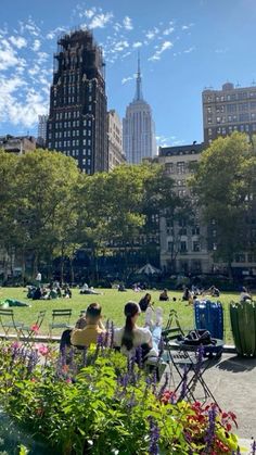 two people sitting on a bench in the middle of a park with buildings in the background