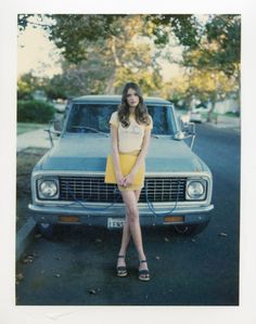 a woman standing in front of a blue car on the street with her legs crossed