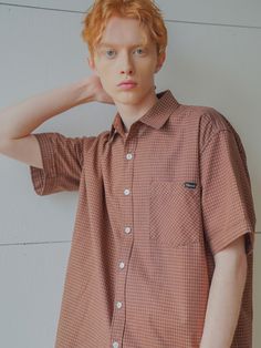 a young man with red hair wearing a brown checkered shirt and blue jeans standing against a white wall