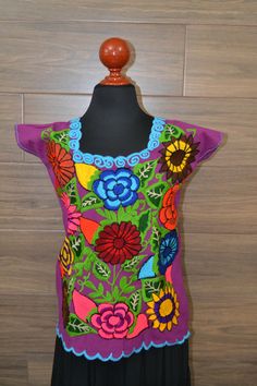 "This handmade Mexican shirt has been embroidered with beautiful colorful flowers. It will look great with any color skirt or pants. It has short sleeves and a scoop neckline. This is a nice comfortable casual cotton shirt. It is perfect as a gift for a friend or someone special. This shirt is a medium size. It measures 19\" wide x 23\" long. It has 7\" sleeve openings and a 8\" opening for the neck. Size Medium About Our Items: Each one of our items is handmade/hand woven by Indigenous communit Purple Embroidered Short Sleeve Blouse, Casual Purple Floral Embroidered Top, Casual Purple Top With Floral Embroidery, Casual Purple Embroidered Top, Embroidered Bohemian Purple Blouse, Bohemian Embroidered Purple Blouse, Traditional Multicolor Embroidered Floral Top, Purple Embroidered Bohemian Blouse, Traditional Multicolor Embroidered Blouse