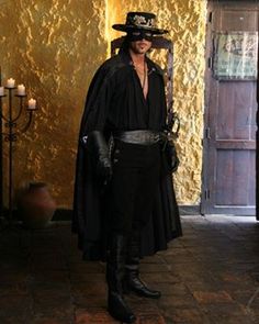 a man dressed in black with a hat and cape standing next to a wall holding a lit candle