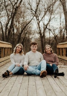 three people sitting on the ground in front of some trees and one person is wearing jeans