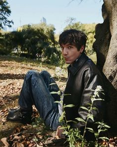 a young man sitting on the ground next to a tree and some leaves in front of him