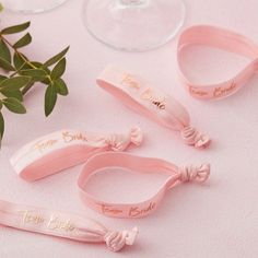 three pink hair ties with names on them sitting next to some wine glasses and greenery