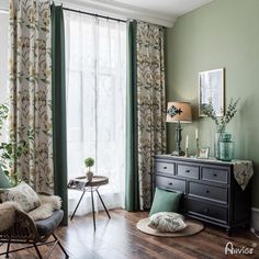 a living room with green walls and curtains, wooden flooring, two wicker chairs and a black dresser