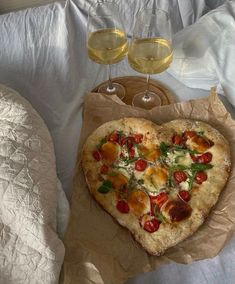 a heart shaped pizza sitting on top of a bed next to two glasses of wine