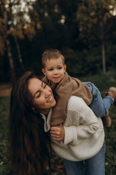 a woman holding a young boy in her arms and smiling at the camera with trees behind her