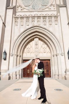 a bride and groom kissing in front of an ornate building with a veil blowing in the wind