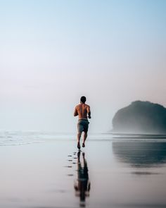 a man running on the beach in front of an ocean with mountains and fog behind him