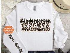 An adorable Halloween sweatshirt or t-shirt for the kindergarten teacher. Customize by adding a name on the sleeve.  Great to wear while teaching.  SWEATSHIRT PRODUCT DESCRIPTION: Ideal for any situation, a unisex heavy blend crewneck sweatshirt is pure comfort. These garments are made from polyester and cotton. This combination helps designs come out looking fresh and beautiful. The collar is ribbed knit, so it retains its shape even after washing. There are no itchy side seams on these sweater School Long Sleeve T-shirt With Name Print, Customizable Long Sleeve T-shirt For School Spirit, White T-shirt For School In Fall, White Sweatshirt For Teacher Appreciation In Fall, Customizable Tops For School Spirit In Fall, Fun Long Sleeve T-shirt With Name Print, Long Sleeve T-shirt For School In Fall, Long Sleeve White T-shirt For Teacher Appreciation, Teacher Halloween