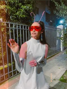 a woman in white dress and red sunglasses standing next to a fence with her hands out