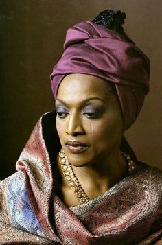 a woman wearing a purple turban and gold jewelry looks off to the side