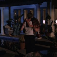 two people standing in front of a house at night with their arms around each other