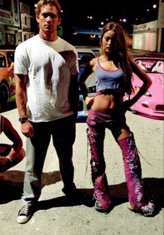 two people standing next to each other in front of a pink car and some buildings