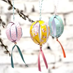 three paper lanterns hanging from a tree branch in front of a white brick wall with flowers