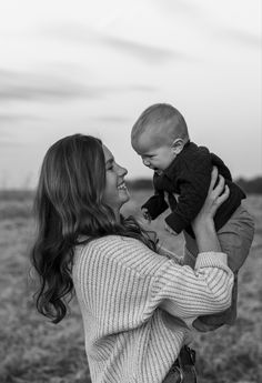 a woman holding a baby up to her face and smiling at the camera while standing in a field