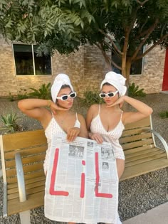 two women in bathing suits sitting on a bench holding up a newspaper with the word lil written on it