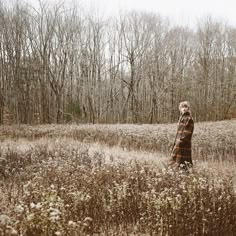 a woman standing in the middle of a field with tall grass and trees behind her