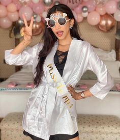 a woman wearing sunglasses and a sash standing in front of a bed with pink balloons