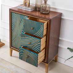 a wooden cabinet with two drawers and an abstract design on the front, next to a potted plant