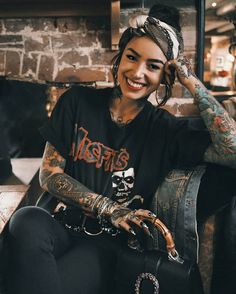 a woman with tattoos sitting in a chair smiling at the camera and holding her hand up to her ear