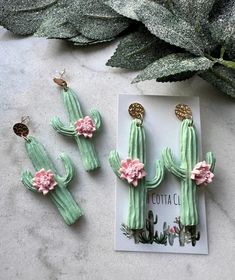 three cactus earrings with pink flowers on them sitting next to a card and green leaves