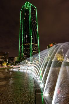 the water fountain is lit up with green lights in front of a tall building at night