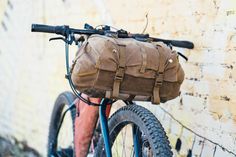 a bicycle with a bag strapped to the front handlebars is parked against a brick wall