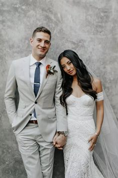 a bride and groom standing next to each other in front of a wall holding hands