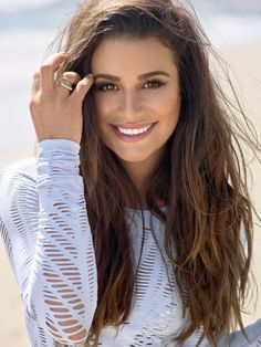 a woman with long hair smiling and holding her hand up to her face while standing on the beach