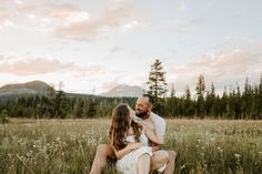 a man and woman sitting on the grass in front of some mountains at sunset with their arms around each other