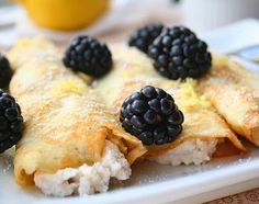 a white plate topped with crepes covered in blackberries and cheese next to a cup of orange juice