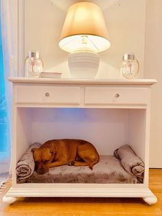 a brown dog laying on top of a bed under a table next to a lamp